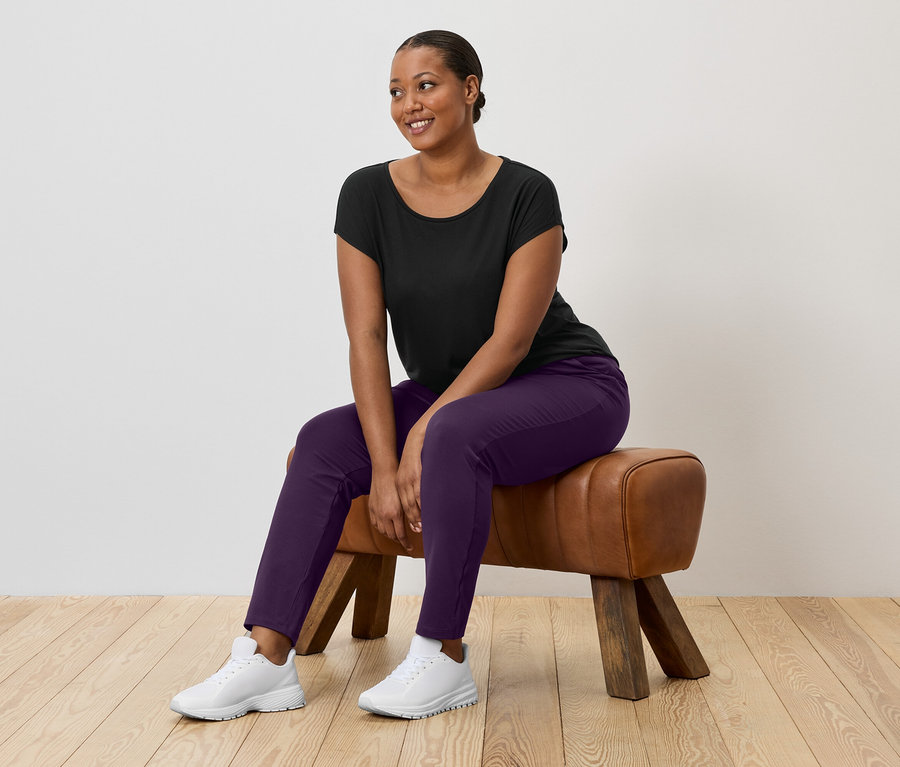 Kobieta o ciemnych włosach siedzi na kozle gimnastycznym, ubrana w czarny t-shirt, fioletowe spodnie i białe buty sportowe.