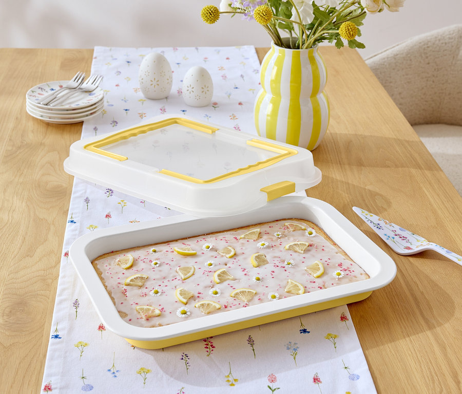 Na drewnianym stole leży ciasto z lukrem cytrynowym i stokrotkami, dzbanek w żółto-białe paski, łopatka do ciasta we wzory kwiatowe i biały obrus we wzory kwiatowe.