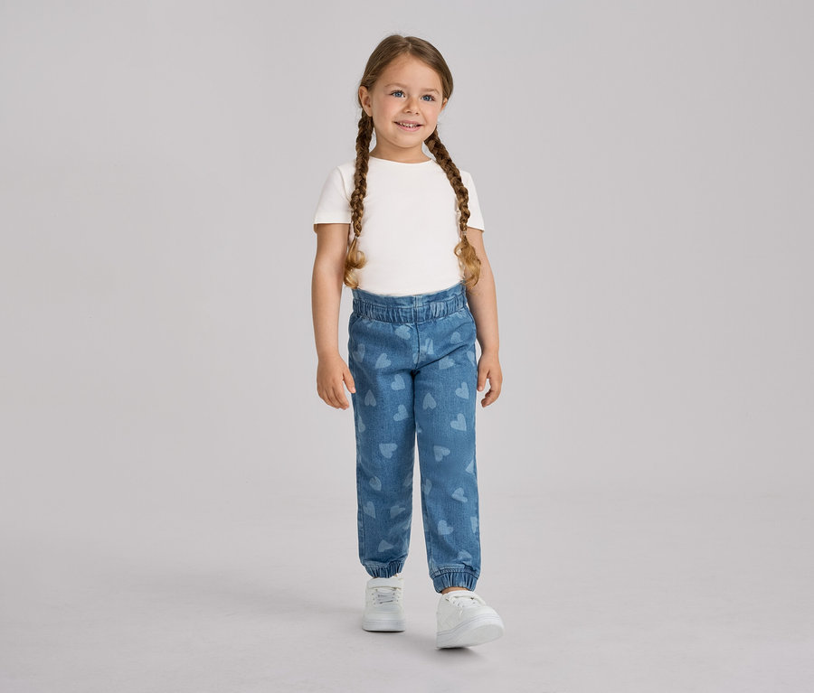 Uśmiechnięta dziewczynka z dwoma warkoczami stoi, ubrana w biały T-shirt, niebieskie spodnie na gumce – fason „Ella” w serduszka i białe trampki.
