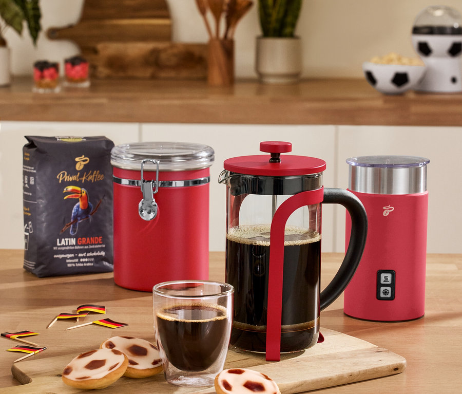 Na stole stoi czerwona puszka zatrzymująca zapach, czerwony elektryczny spieniacz do mleka, czerwony zaparzacz tłokowy, 2 szklanki o podwójnych ściankach do caffè crema i paczka kawy.