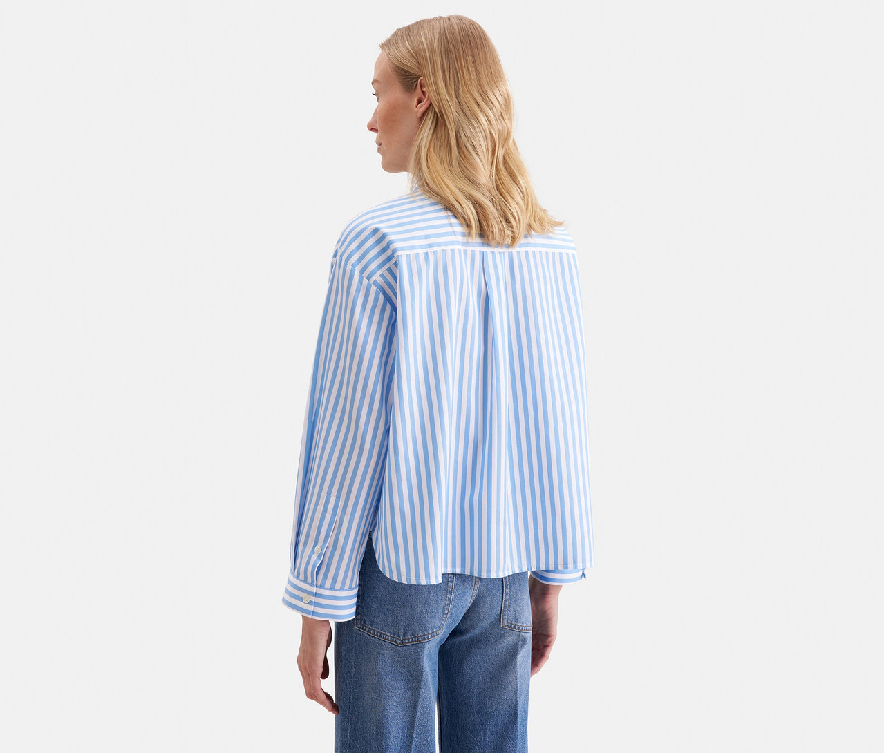 Kobieta z blond włosami, widziana od tyłu, ma na sobie niebiesko-białą bluzkę koszulową oversize Seidensticker w paski z długimi rękawami i niebieskie dżinsy.