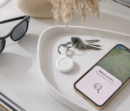 Na tacy leżą okulary przeciwsłoneczne, klucze, biały lokalizator i smartfon z aplikacją map.