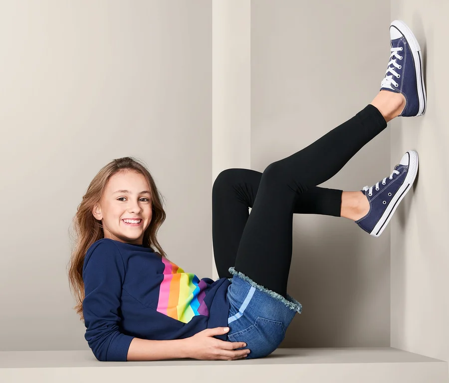Dziewczyna leży i ma na sobie czarne legginsy dziecięce z bawełny ekologicznej (2 pary), dżinsowe szorty i niebieskie trampki.
