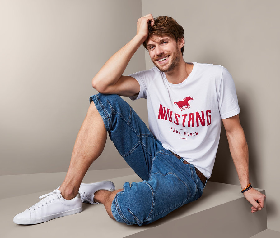 Mężczyzna siedzi i ma na sobie męskie bermudy dżinsowe »Mustang«, model Fremont i t-shirt.