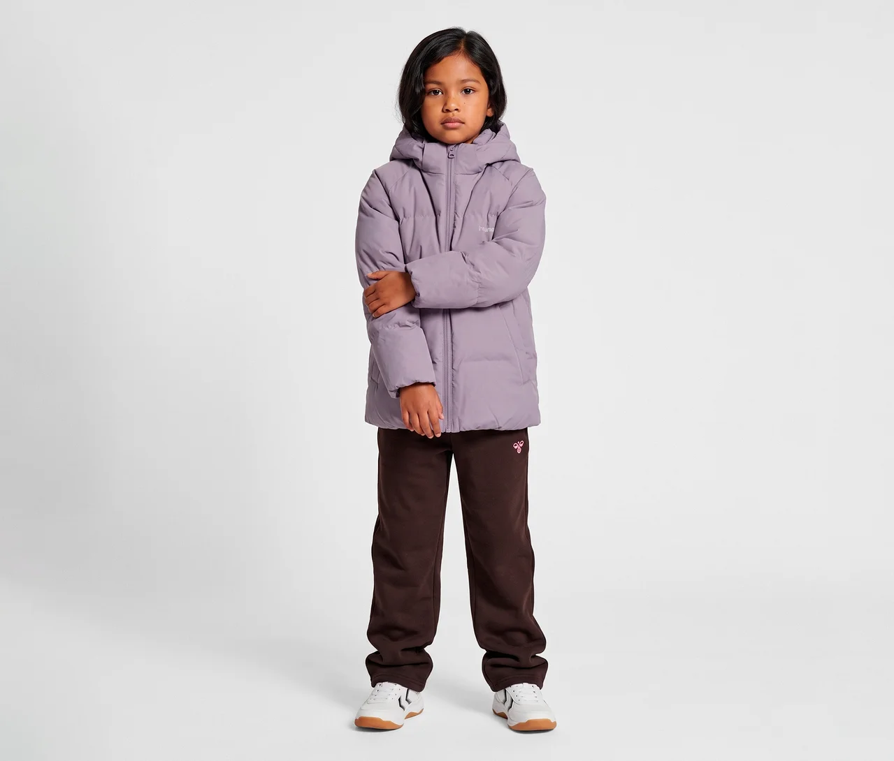 Dziecko z ciemnymi włosami stoi frontalnie, ubrane w fioletową kurtkę puchową hummel® »HMLJR All Weather Puffer Jacket« z kapturem, ciemnobrązowe spodnie dresowe i białe sneakersy. Ręce dziecka są skrzyżowane.