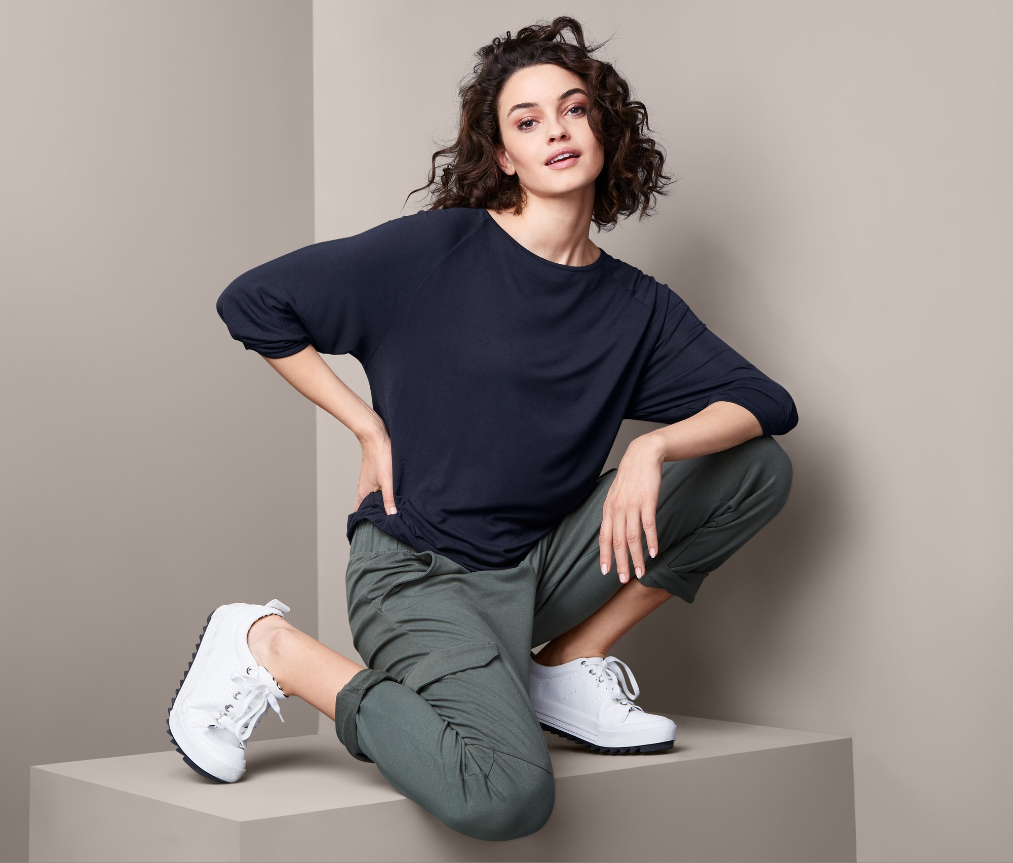 Kobieta w czarnej bluzce z kimonowymi rękawami i zielonych spodniach kuca.