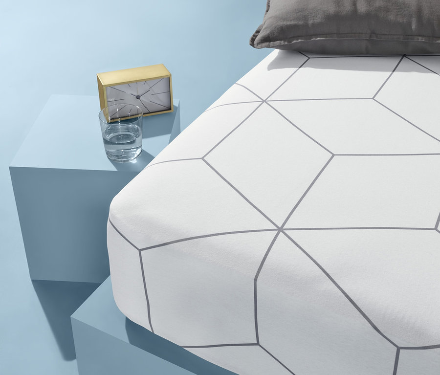 Na niebieskim tle stoi łóżko z białą pościelą w geometryczny wzór i szarą poduszką. Obok łóżka na niebieskim stoliku stoi złoty zegar i szklanka wody.