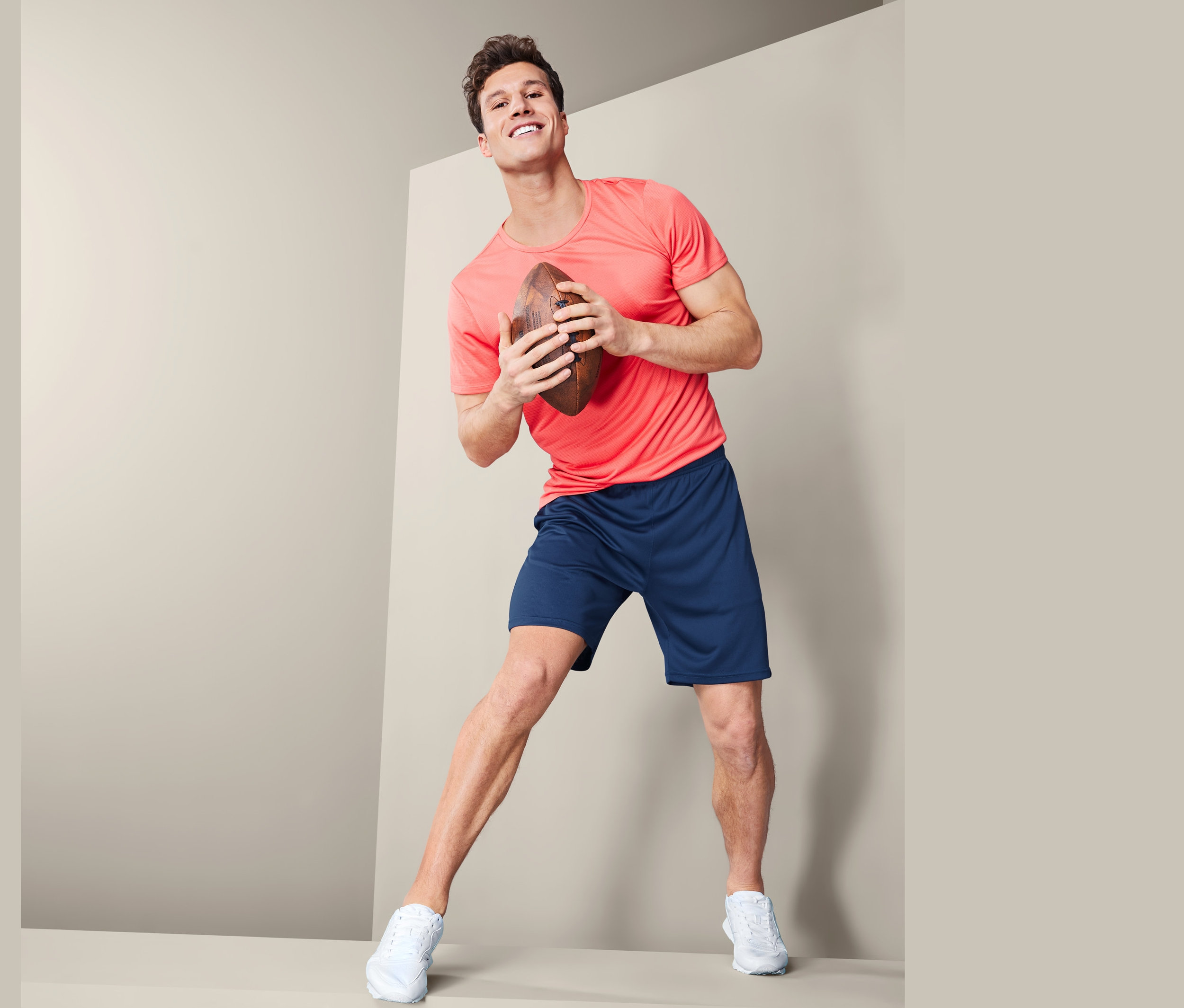 Mężczyzna trzyma piłkę do futbolu amerykańskiego i ma na sobie męskie szorty sportowe, granatowe.