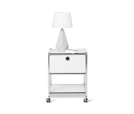 Na białym tle biały metalowy stolik »CN3« z szufladą, na którym stoi lampa i leży książka.
