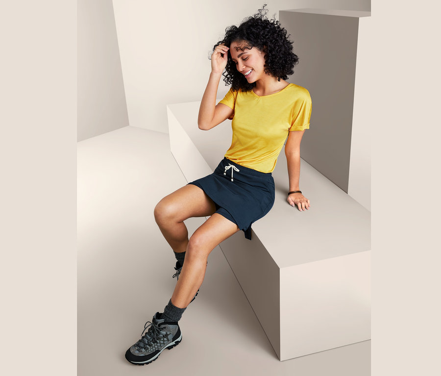 Kobieta siedzi i ma na sobie żółtą koszulkę i granatowe szorty ze spódniczką.