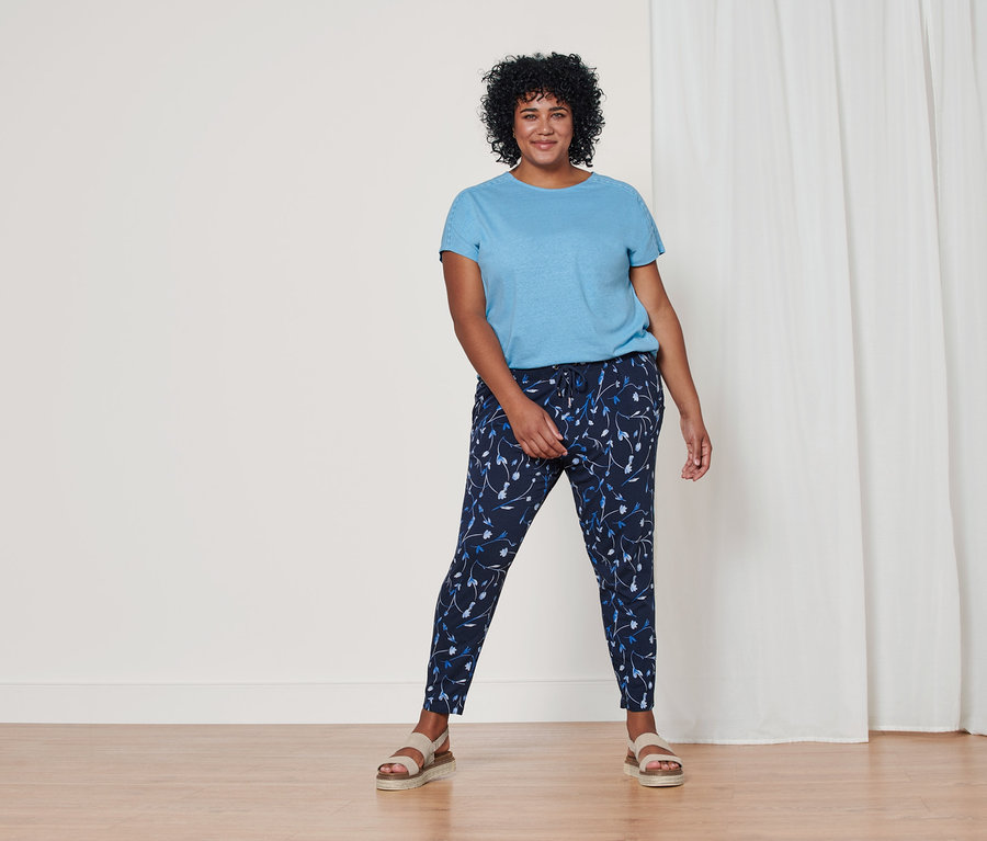 Kobieta stoi ubrana w błękitną bluzkę z mieszanki materiałowej z zawartością lnu, granatowe spodnie dżersejowe z nadrukiem na całej powierzchni i skórzane sandały na platformie.