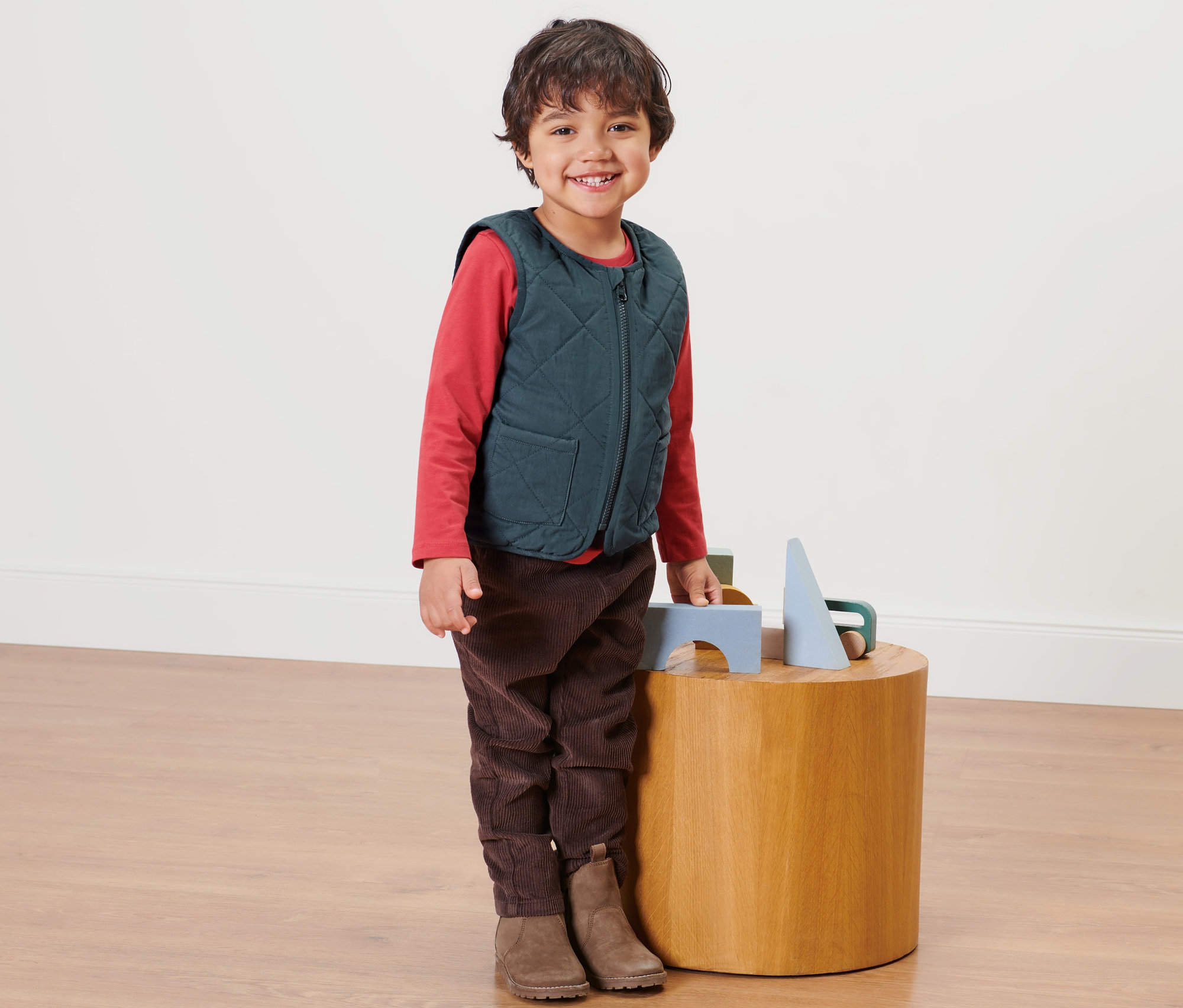 Chłopiec stoi obok drewnianego stolika i ma na sobie dziecięcą pikowaną kamizelkę.