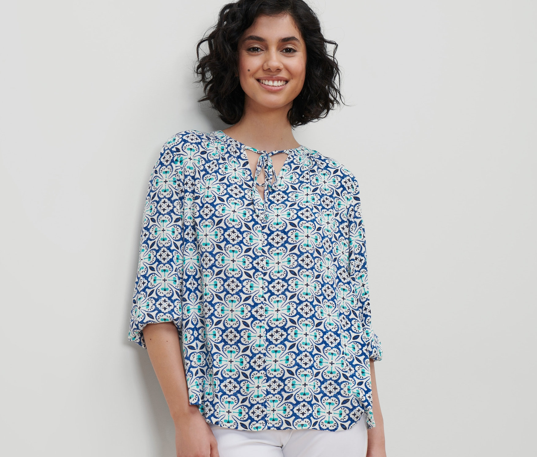 Kobieta ma na sobie bluzkę-tunikę z wzorem na całej powierzchni i białe spodnie.