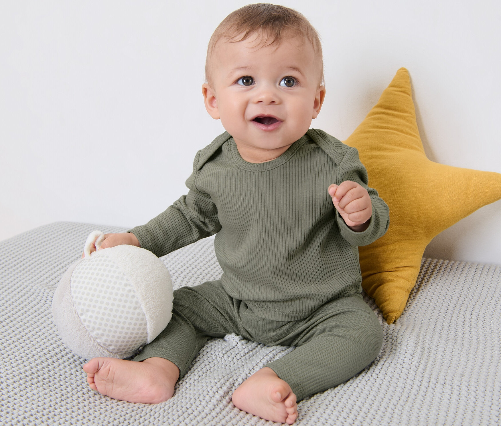 Dziecko siedzi na szarym kocu obok żółtej gwiazdy. Ubrane jest w koszulkę trykotową z zestawu 3 sztuk.