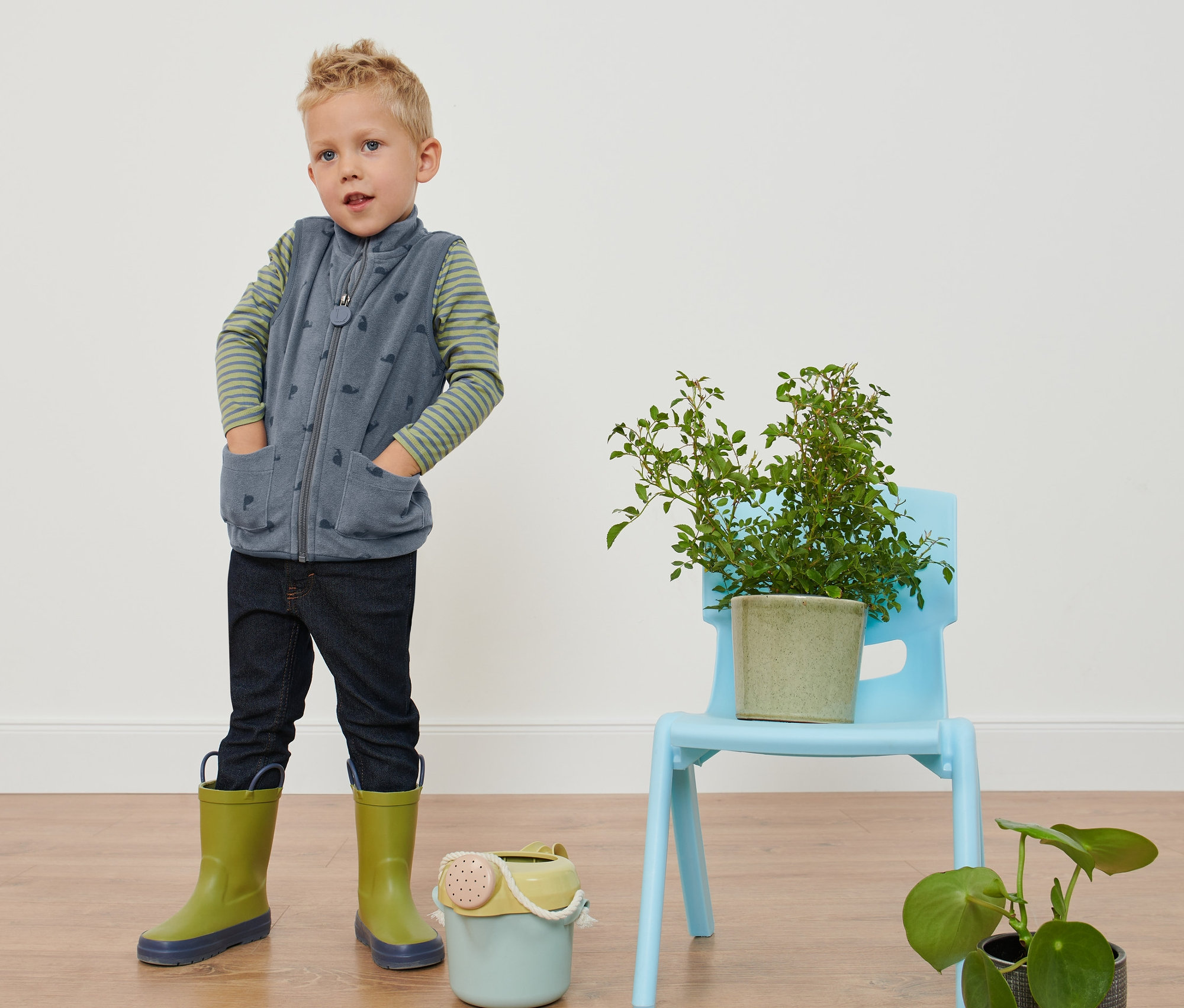 Chłopiec stoi z rękami w kieszeniach w kamizelce z polaru w ślimaki, dżinsach i kaloszach.