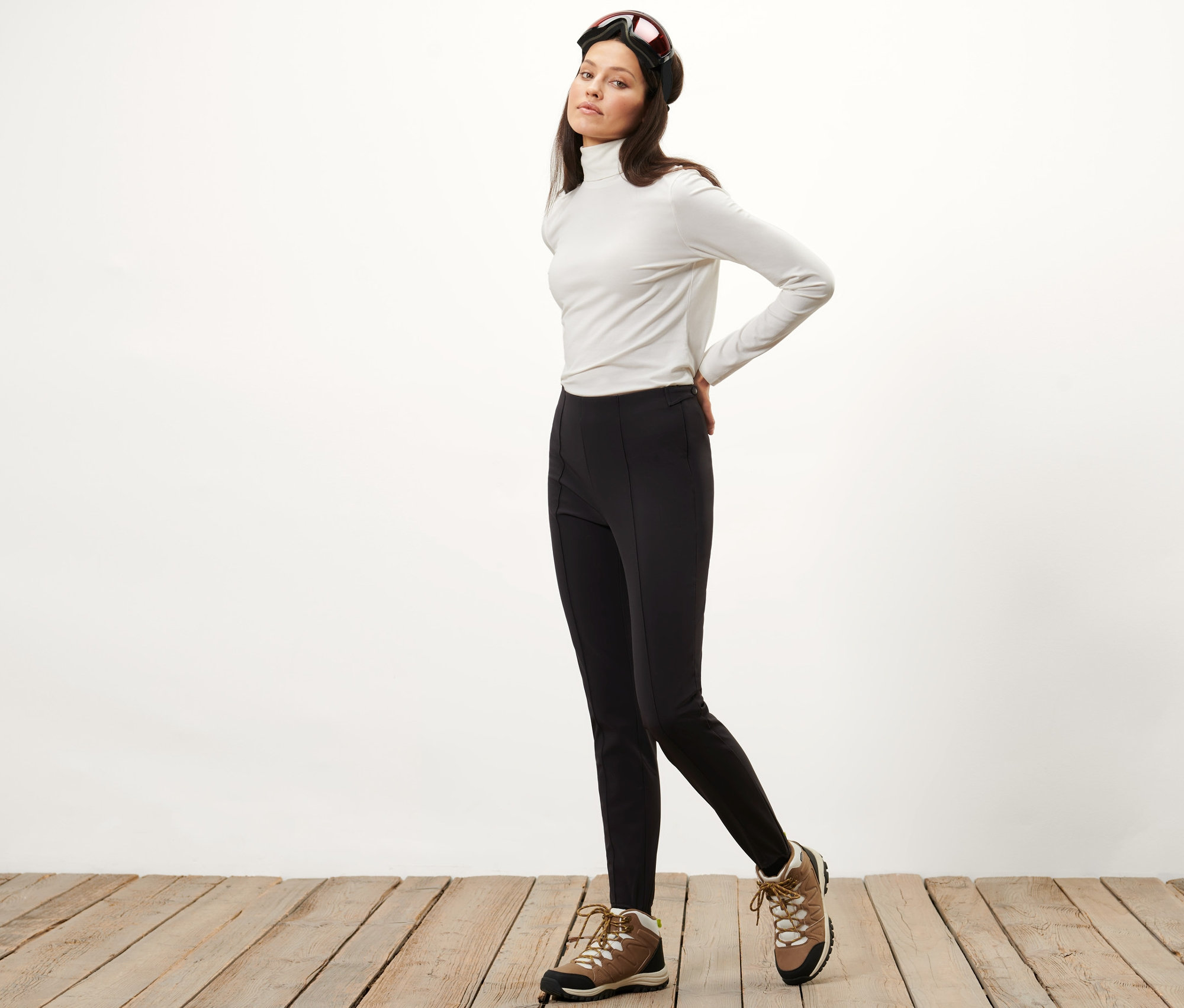 Kobieta pozuje w narciarskich, obcisłych spodniach z tkaniny softshell, białym golfie i goglach narciarskich.