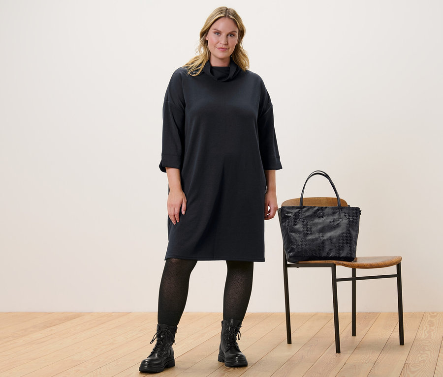 Kobieta stoi i ma na sobie sukienkę dresową z mieszanki modalu oraz rajstopy dzianinowe ze strukturą. Obok niej stoi krzesło, na którym leży torebka.