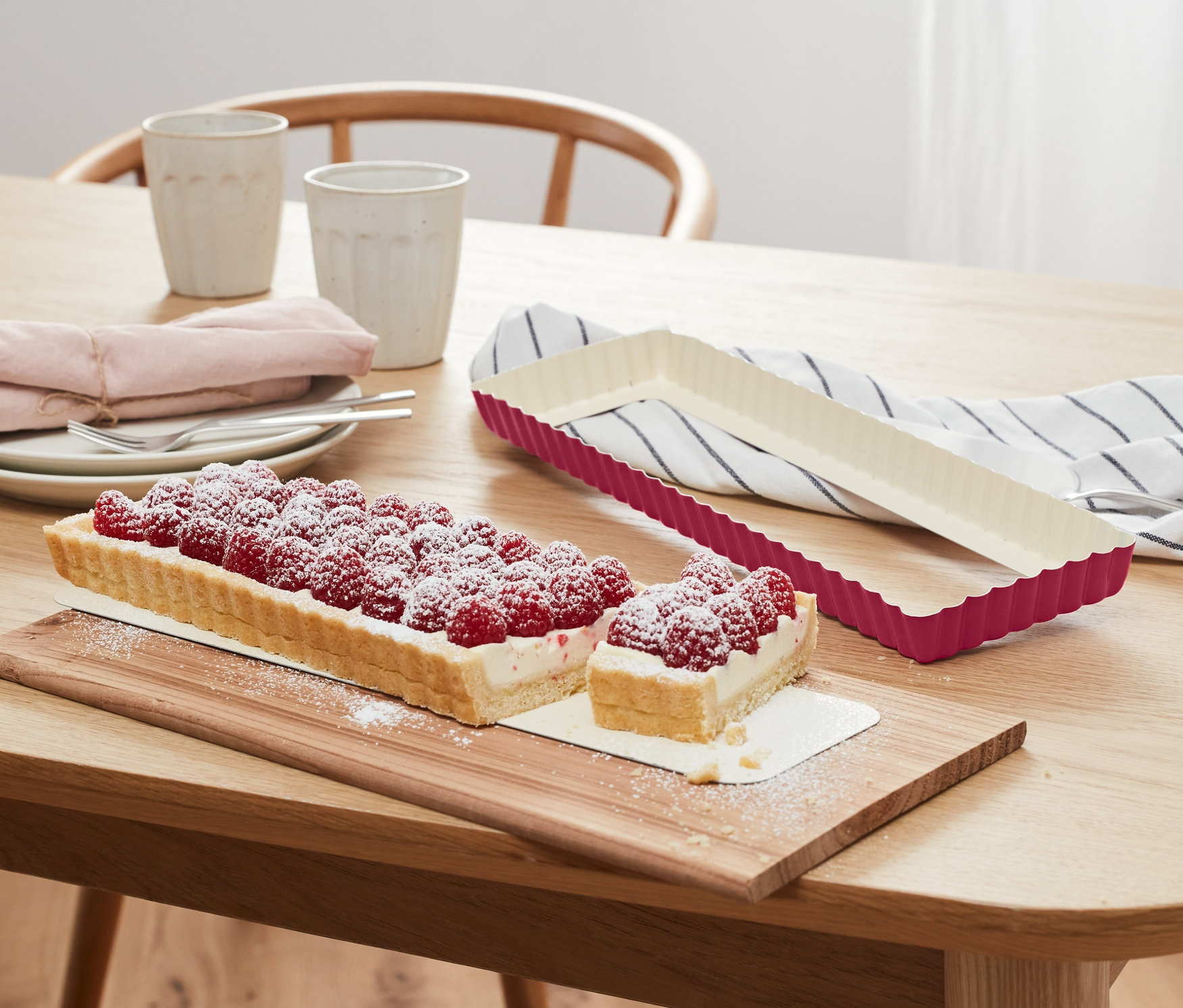 Na drewnianym stole leży forma do wypieku tart i pokrojony torcik malinowy.