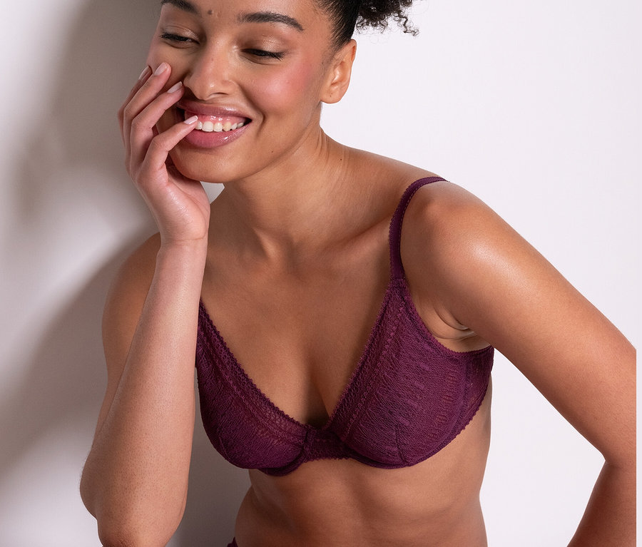 Uśmiechnięta kobieta siedzi i ma na sobie fioletowy biustonosz z fiszbinami Passionata „Leila” i pasujące majtki.