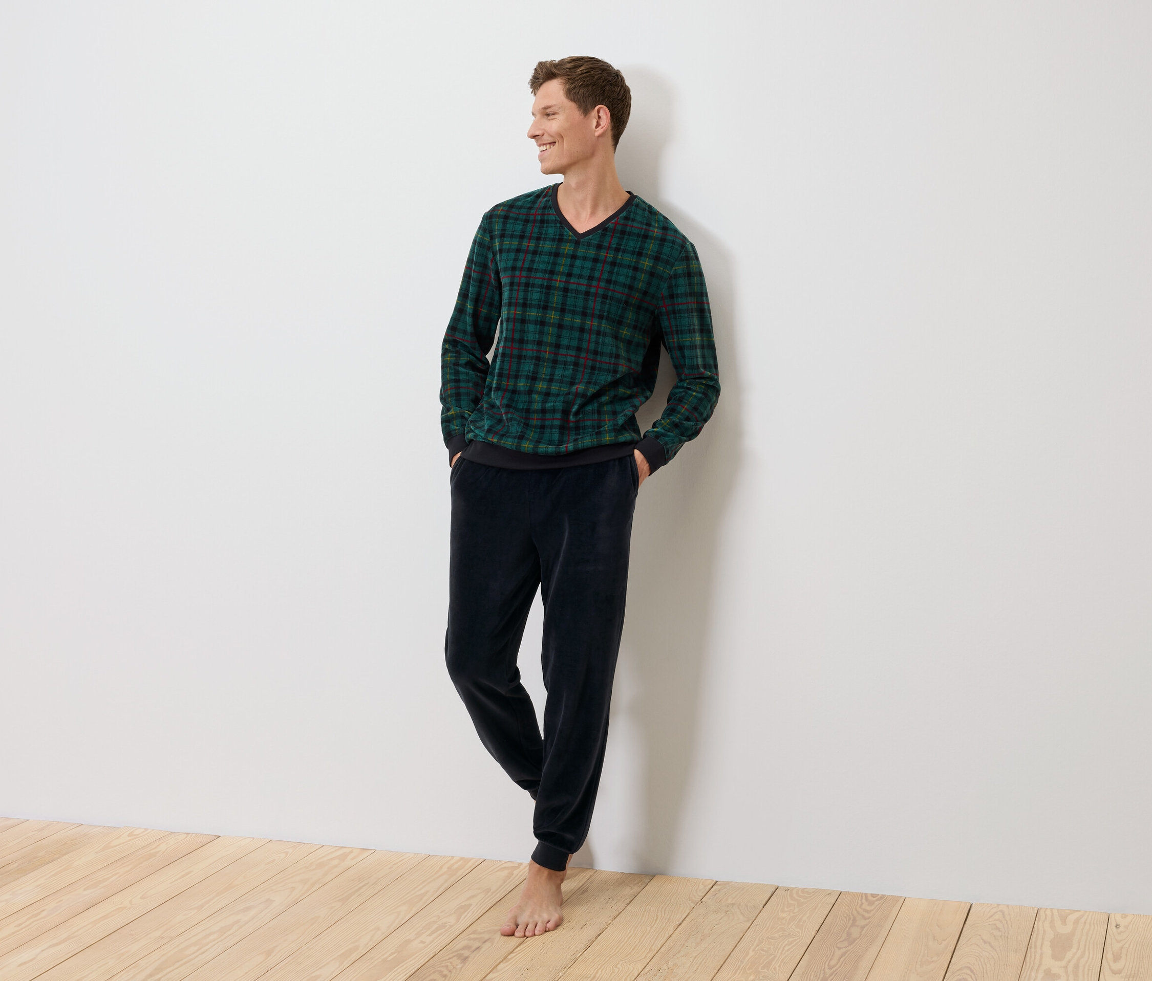 Mężczyzna stoi oparty o ścianę, ubrany w zielony sweter w kratę i granatowe spodnie welurowe.