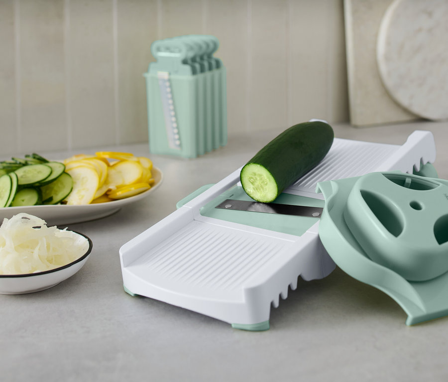 Zielony ogórek na białej krajalnicy do warzyw z akcesoriami. Na talerzu pokrojone ogórki i żółta dynia.