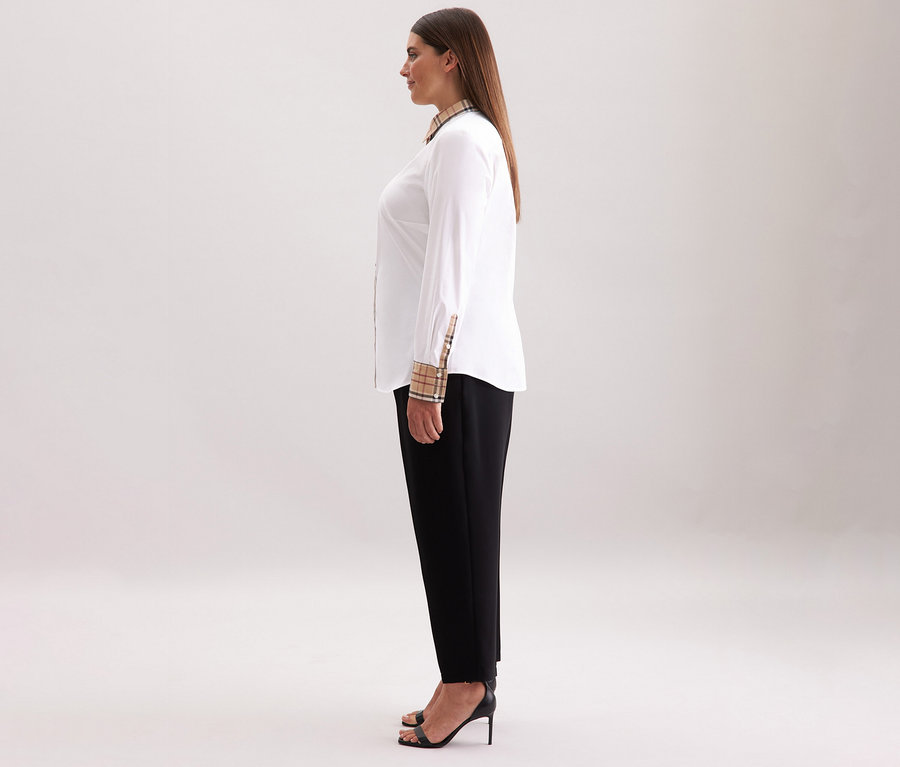 Kobieta stoi bokiem, ubrana w białą popelinową koszulę Seidensticker z kratkowanymi detalami, czarne spodnie i czarne szpilki.