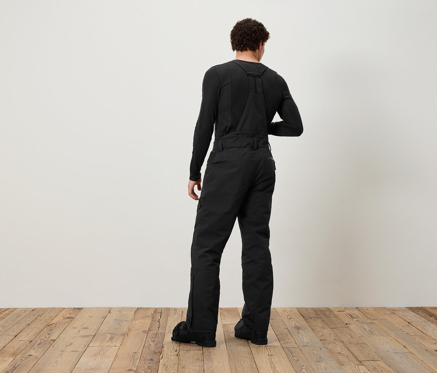 Mężczyzna tyłem do kamery ubrany jest w czarne ogrodniczki i czarną koszulę z długim rękawem.