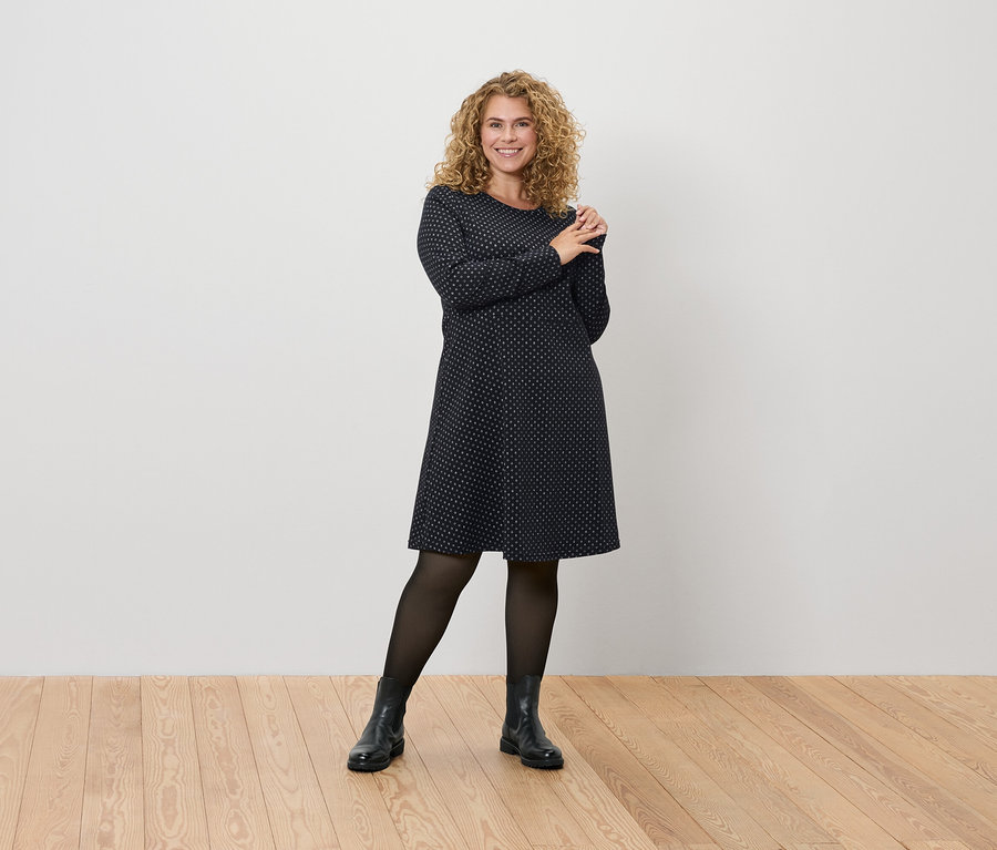 Kobieta pozuje w granatowej sukience w kropki, czarnych rajstopach i butach.