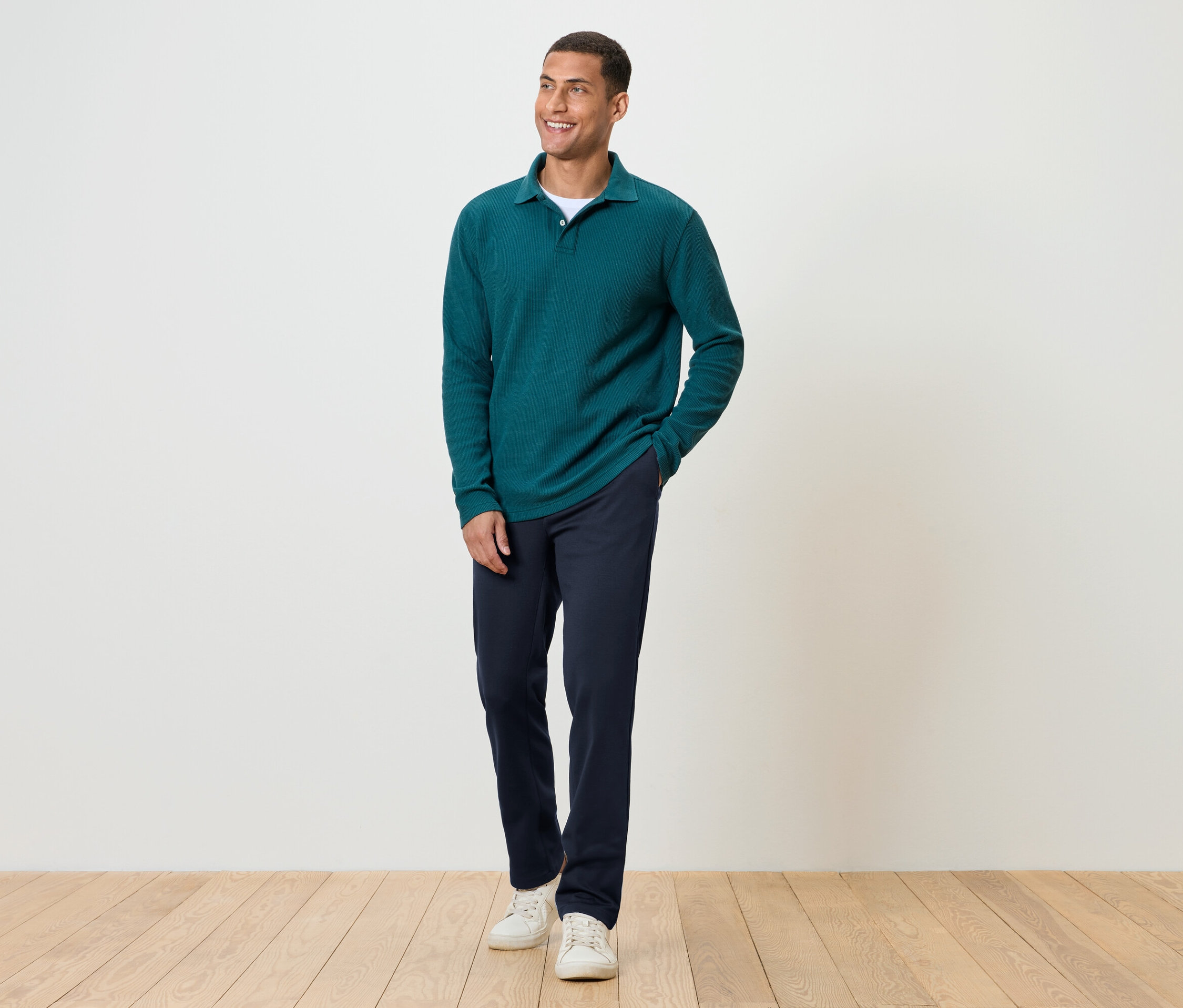 Mężczyzna stoi na drewnianej podłodze, ubrany w granatowe spodnie, zieloną koszulkę z długim rękawem i białe tenisówki.