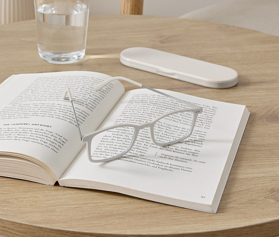 Okulary leżą na otwartej książce na okrągłym stole. Obok książki stoi szklanka wody i etui na okulary.