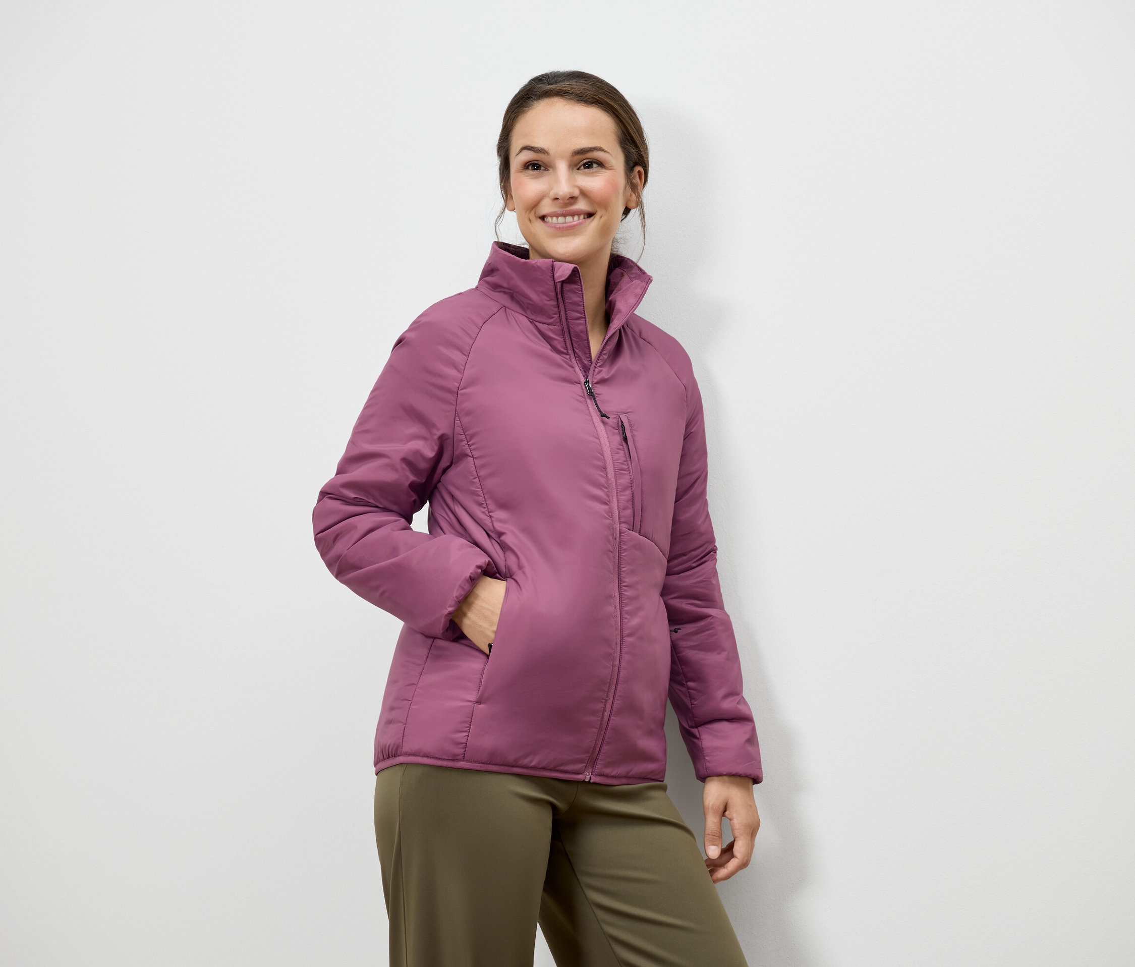 Kobieta z brązowymi włosami i uśmiechem pozuje w różowej kurtce i zielonych spodniach na tle białej ściany.