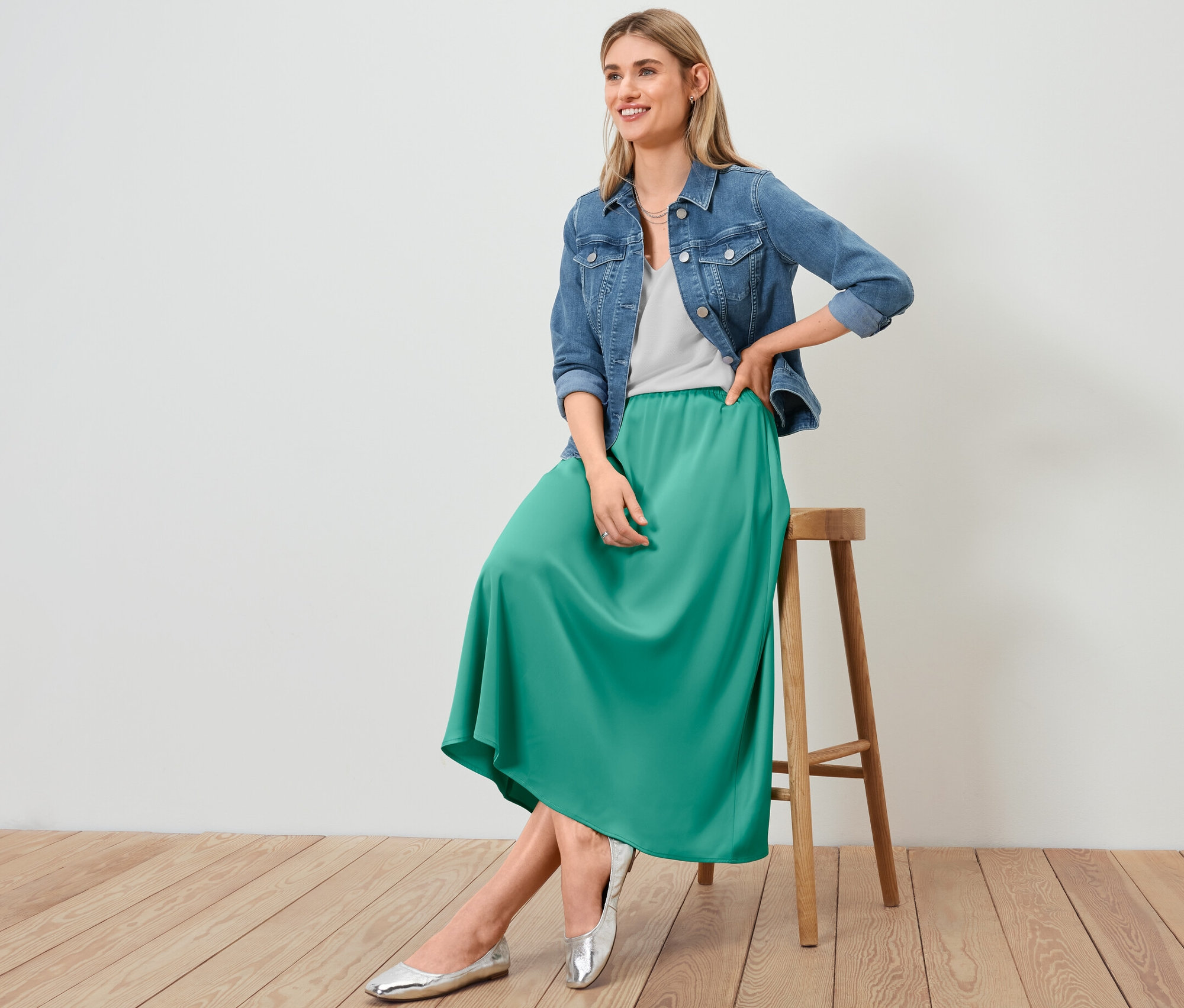 Kobieta siedzi na stołku ubrana w kurtkę dżinsową, spódnicę z satyny i białą koszulkę z fakturowaną powierzchnią.