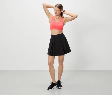 Kobieta z podniesionymi rękami ma na sobie koralowy top sportowy, czarną spódnicę i czarne tenisówki.