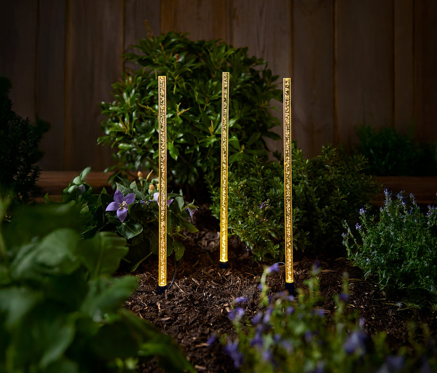 Trzy złote solarne lampy ogrodowe stoją na rabacie z krzewami i kwiatami.