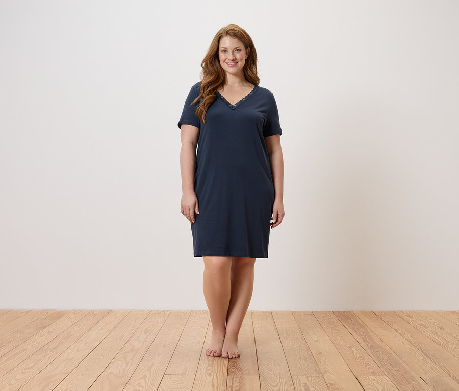Kobieta stoi boso na drewnianej podłodze i ma na sobie granatową koszulę nocną.
