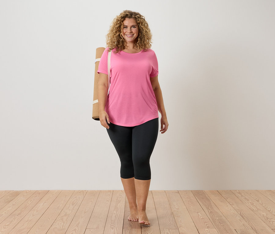 Kobieta z kręconymi włosami ma na sobie różowy T-shirt i czarne legginsy, z matą do jogi na ramieniu, boso na drewnianej podłodze.