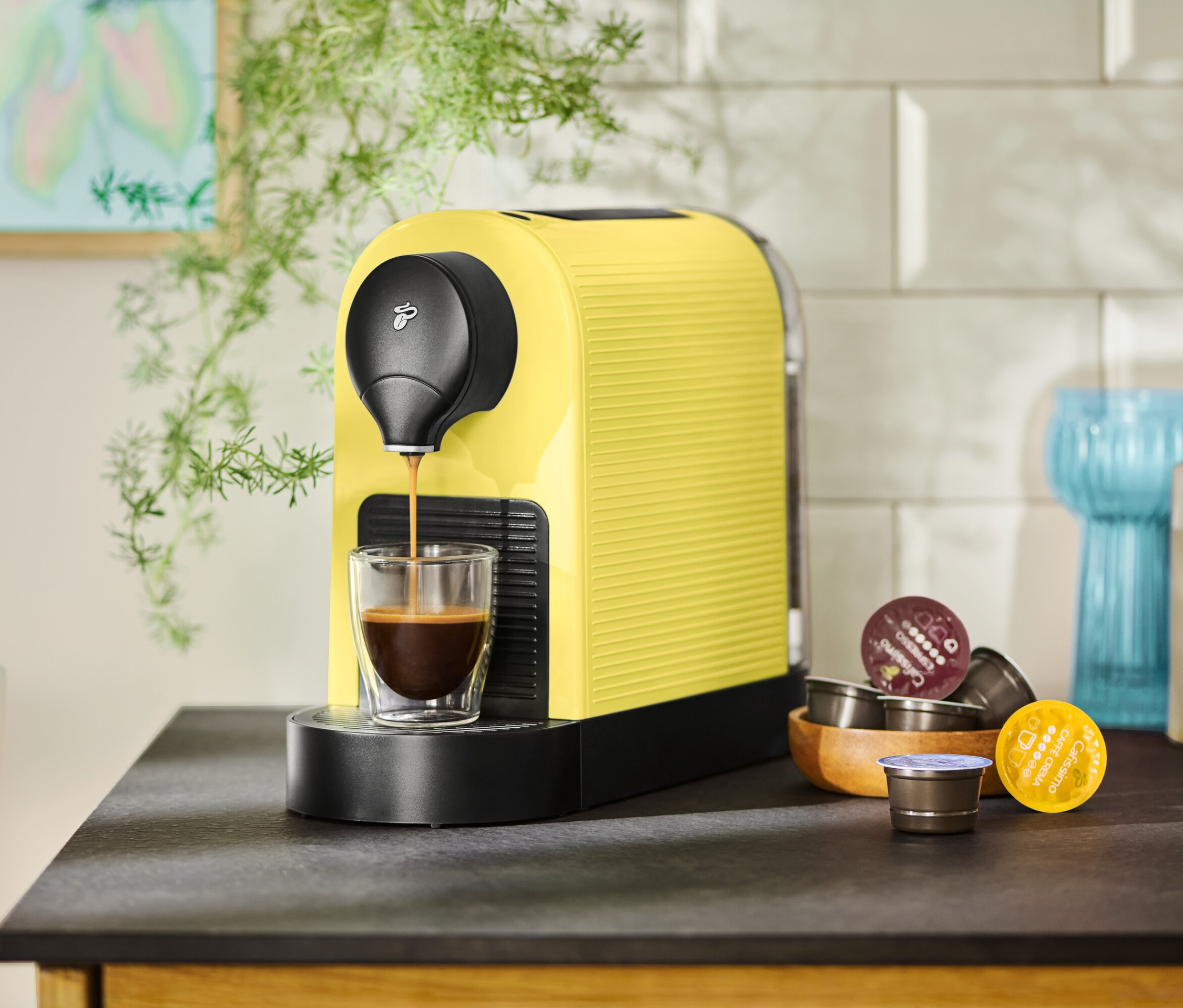 Żółty ekspres do kawy napełnia filiżankę kawą, obok leżą kapsułki.