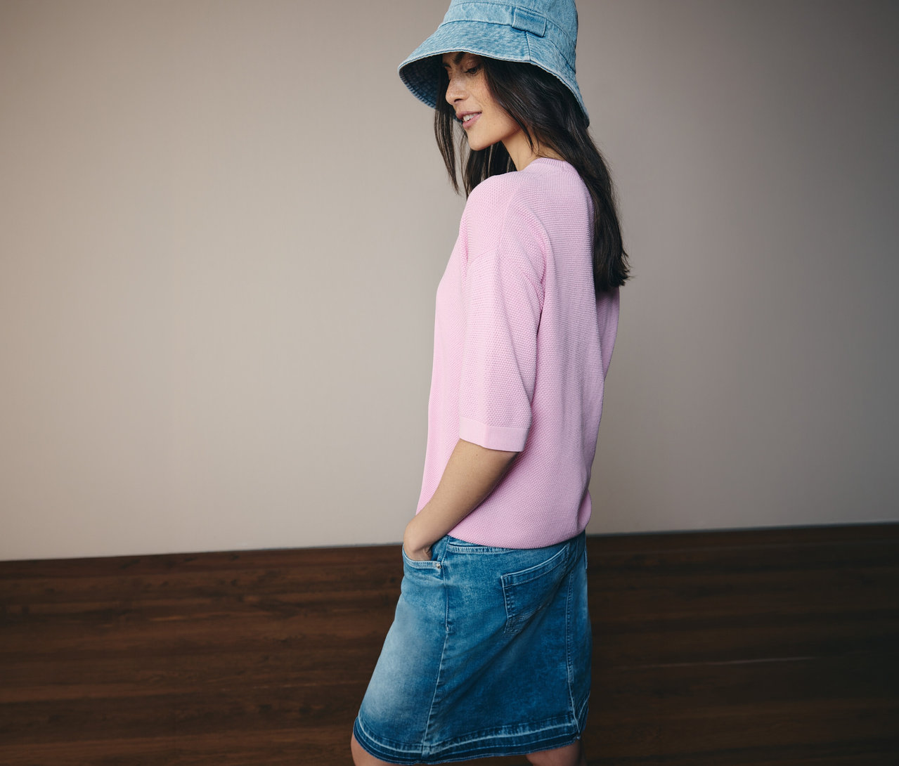 Kobieta w dżinsowym kapeluszu, różowym swetrze z krótkim rękawem CECIL i spódnicy dżinsowej CECIL. Ręce ma w kieszeniach.