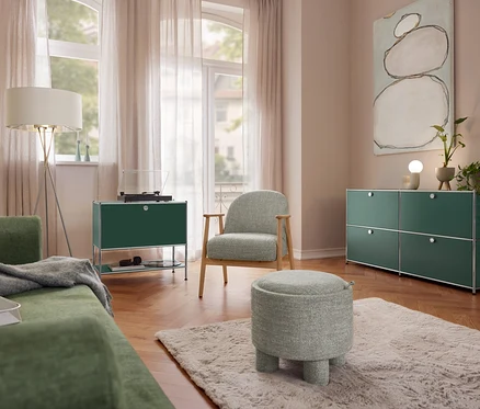 Salon z zieloną sofą, lampą stojącą Eglo „Fondachelli”, metalową szafką typu sideboard „CN3” z 1 klapą i gramofonem. Fotel na drewnianej ramie i pufa do siedzenia stoją na dywanie. Po prawej metalowa szafka typu sideboard „CN3” z 4 klapami i ceramiczna lampa stołowa Trio.