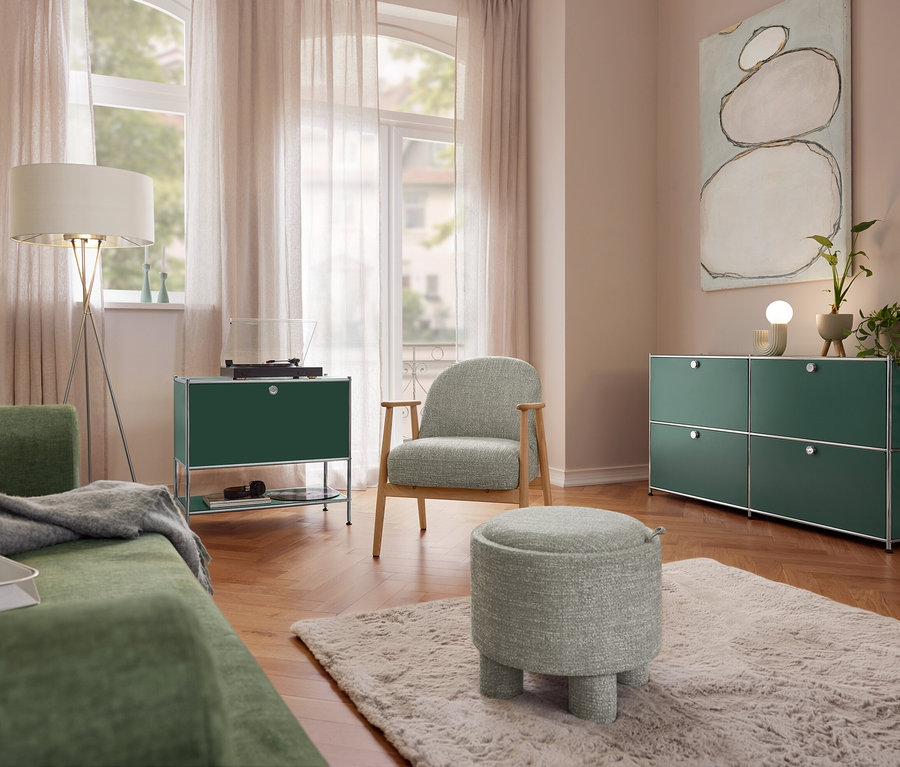 Salon z zieloną sofą, lampą stojącą Eglo „Fondachelli”, metalową szafką typu sideboard „CN3” z 1 klapą i gramofonem. Fotel na drewnianej ramie i pufa do siedzenia stoją na dywanie. Po prawej metalowa szafka typu sideboard „CN3” z 4 klapami i ceramiczna lampa stołowa Trio.