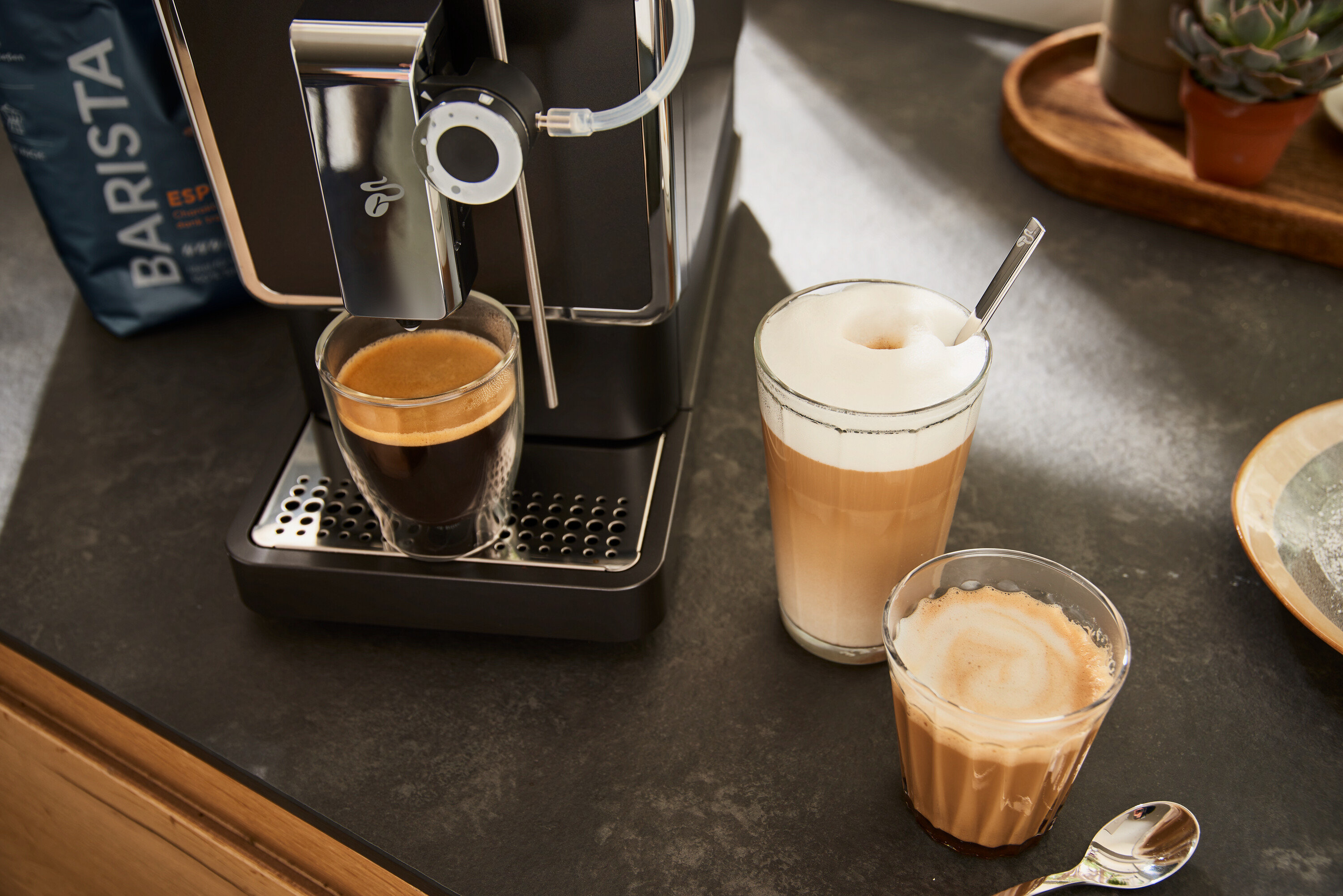 Antracytowy automatyczny ekspres do kawy Tchibo »Esperto Pro« przygotowuje kawę do szklanki. Obok stoją szklanka latte macchiato i szklanka espresso macchiato.