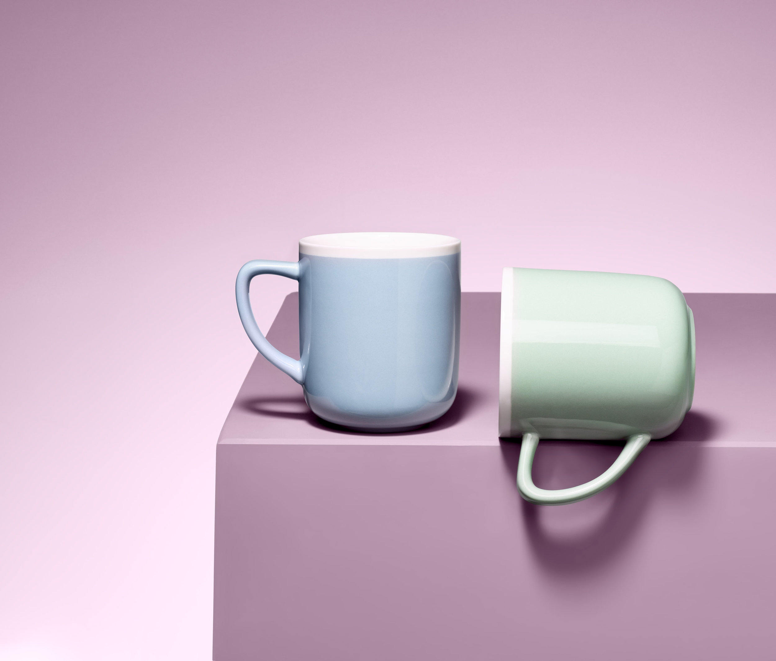 Dwa ceramiczne kubki po 350 ml na fioletowej powierzchni. Jeden jasnoniebieski, drugi jasnozielony.