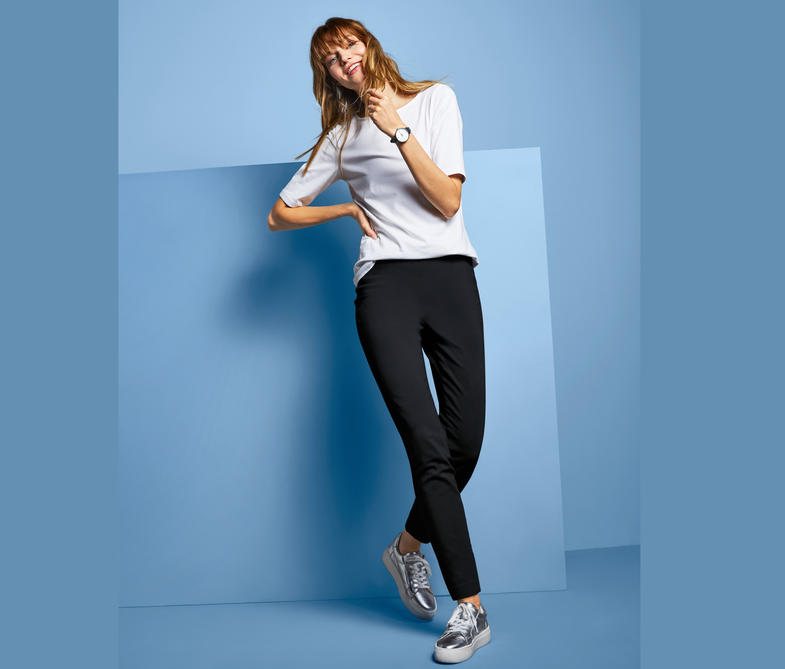 Kobieta pozuje w czarnych spodniach ze stretchu z wąskimi nogawkami, białym t-shircie i srebrnych butach.