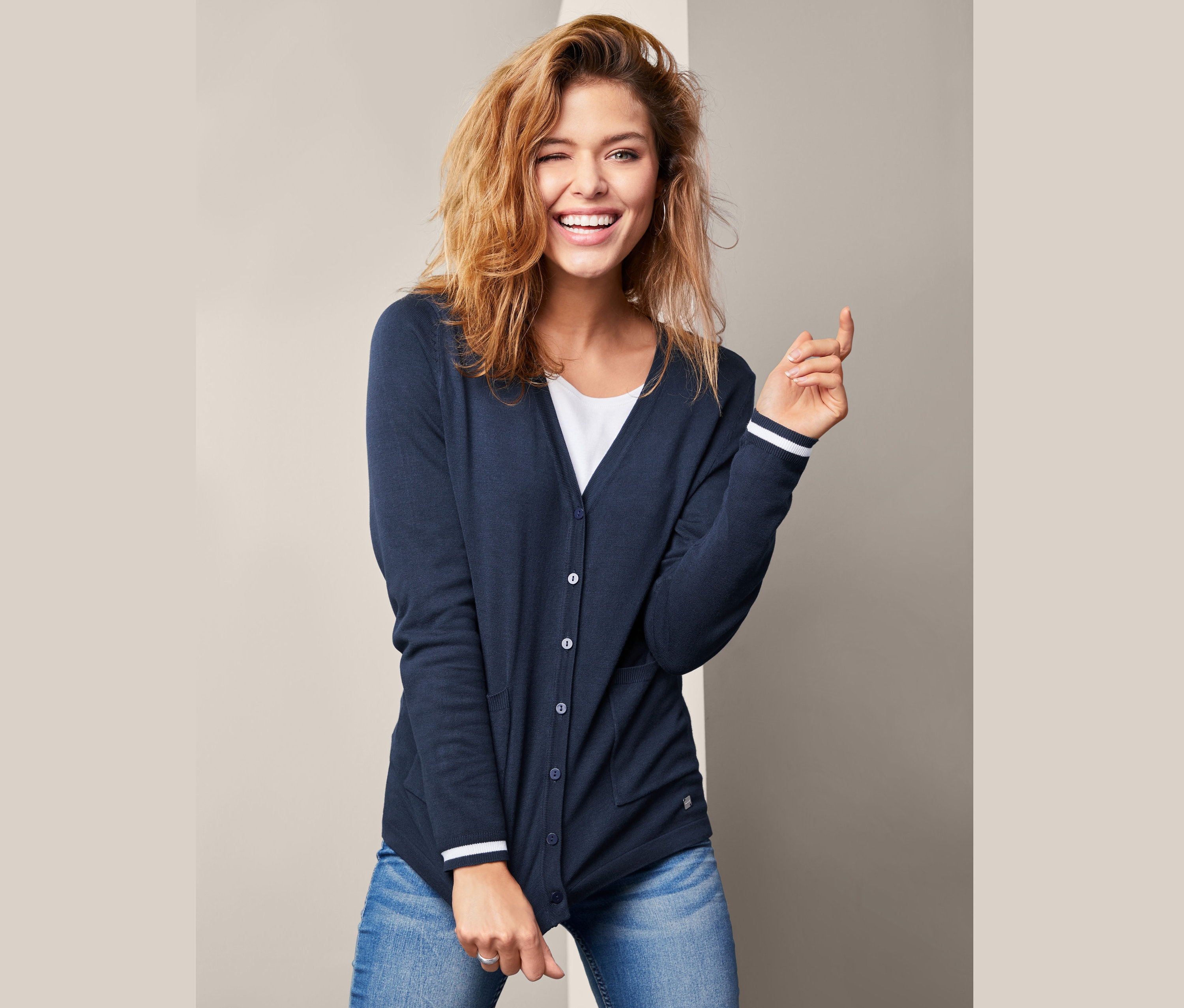 Uśmiechnięta kobieta mruga okiem, ubrana w granatowy damski rozpinany sweter z dekoltem w serek i dżinsy.