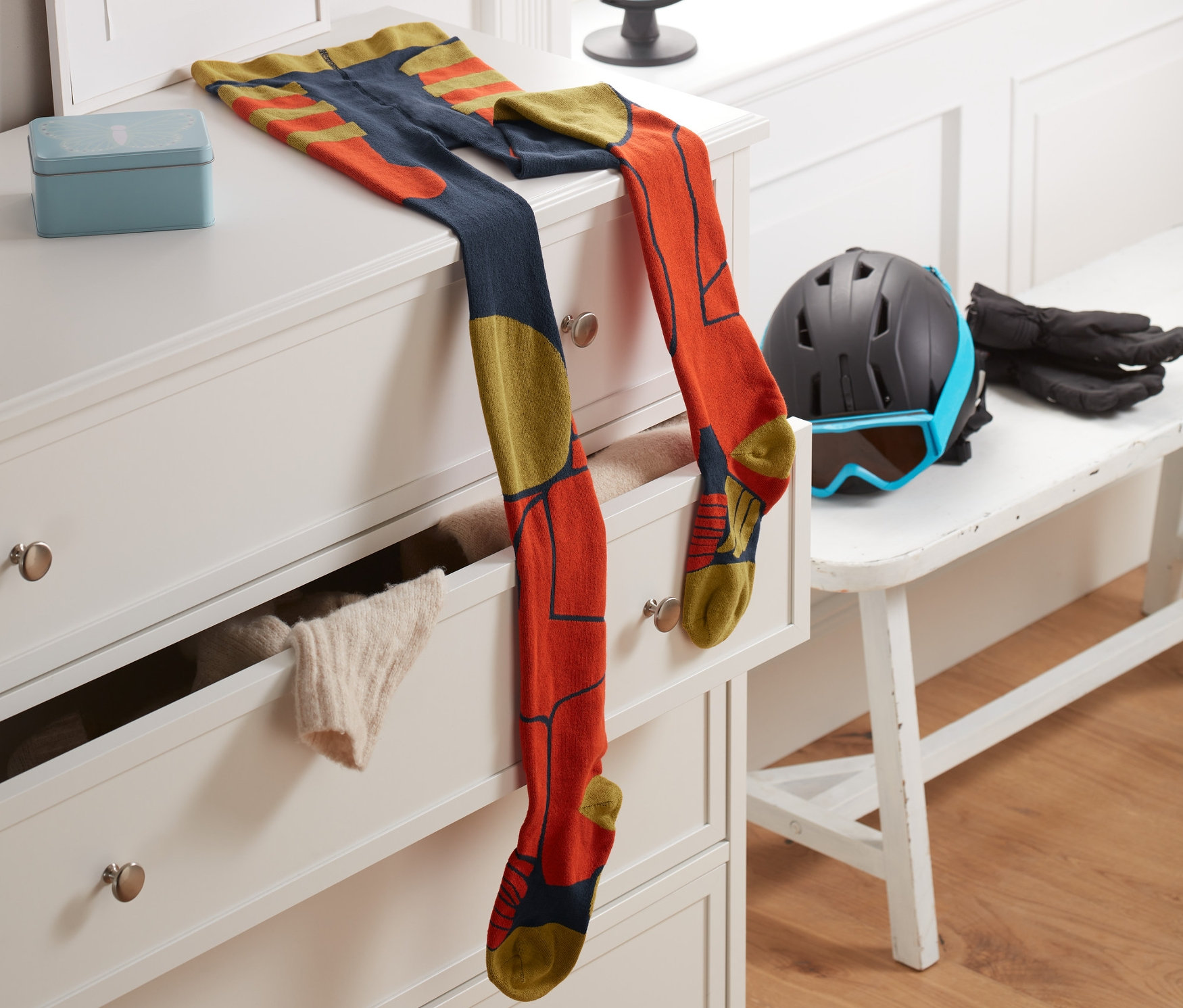 Rajstopy narciarskie leżą na białej komodzie obok ramki z literą T. W tle widoczne okno i globus.