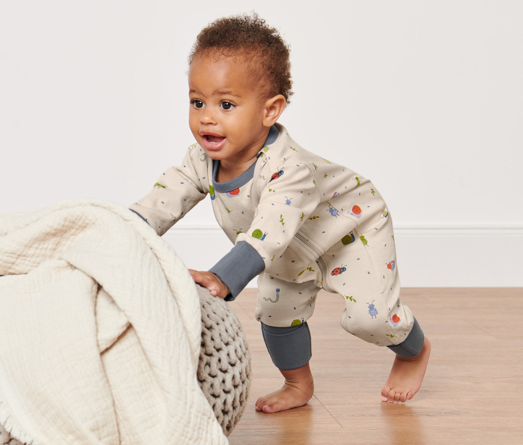 Raczkujące dziecko w piżamie zapinanej na zamek błyskawiczny opiera się o puf z narzuconym kocem.