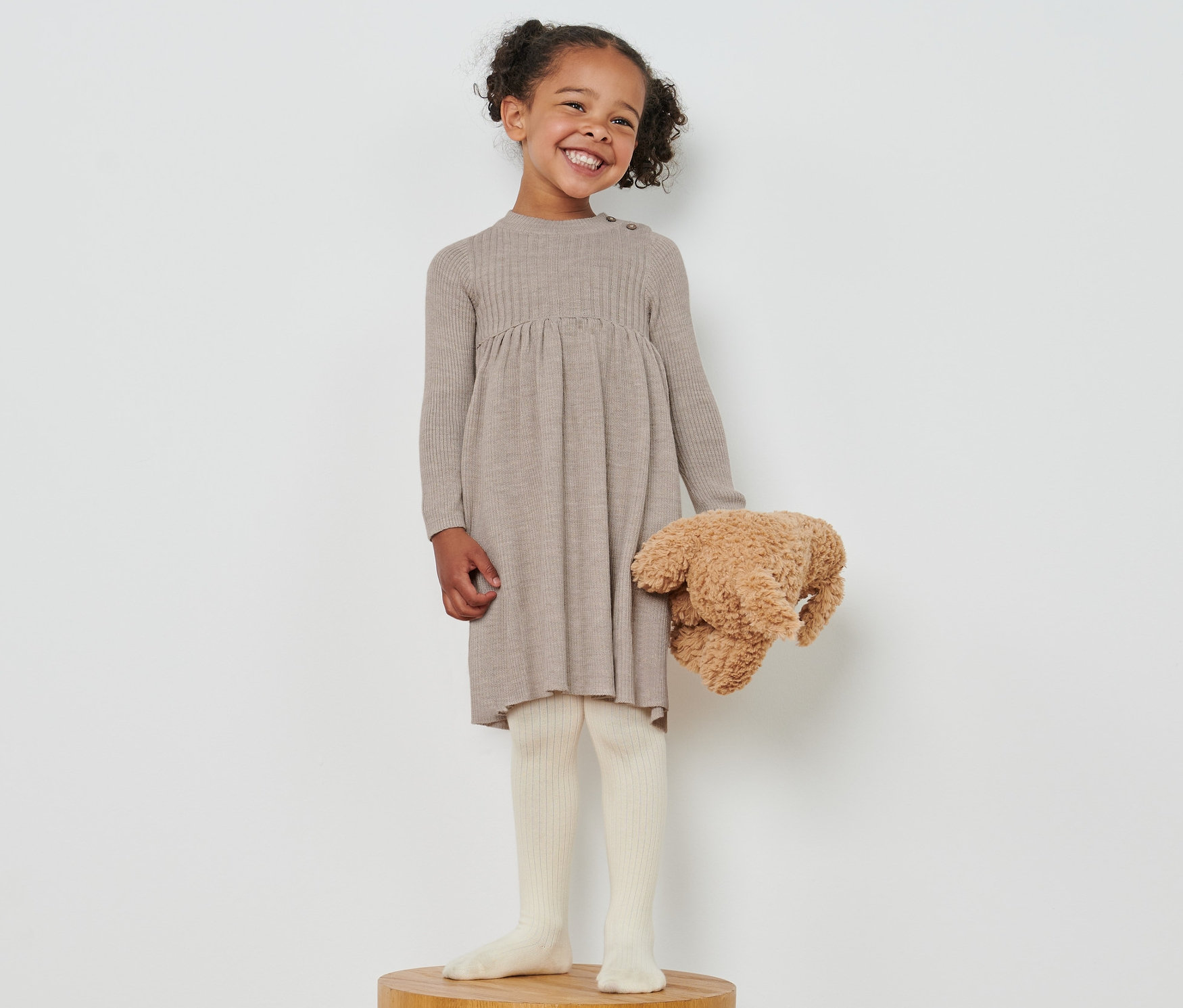 Dziewczynka stoi na drewnianym postumencie, trzyma pluszowego misia i ma na sobie sukienkę z wełny z certyfikatem RWS.