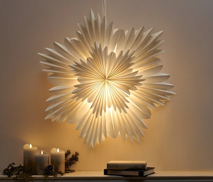 Wisząca papierowa gwiazda LED, XL, świeci nad trzema świecami i dwiema książkami na gzymsie.