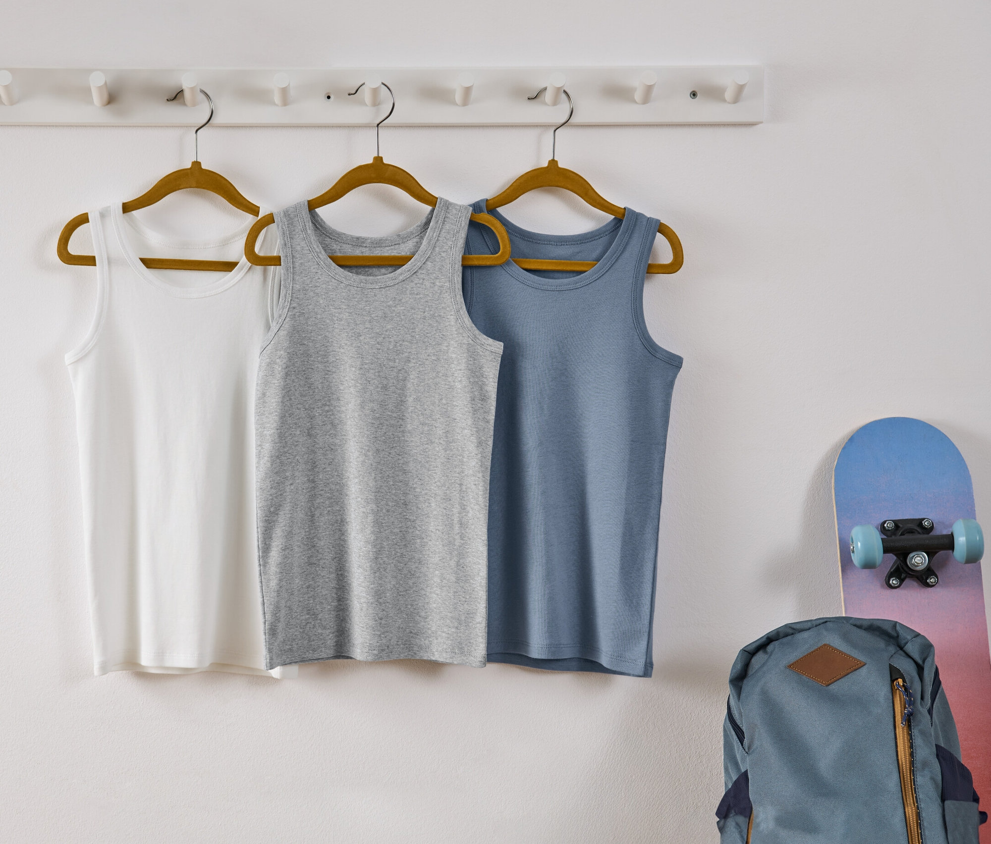 Trzy podkoszulki dziecięce wiszą na wieszakach na białej ścianie. Podkoszulki są w kolorach białym, szarym i niebieskim.