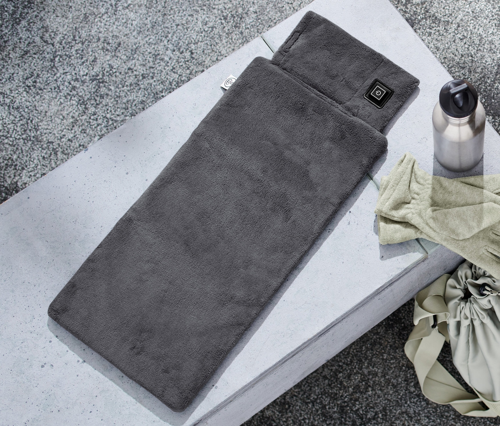 Szara poduszka elektryczna outdoorowa leży na betonowej powierzchni obok butelki z wodą, rękawic i torby.